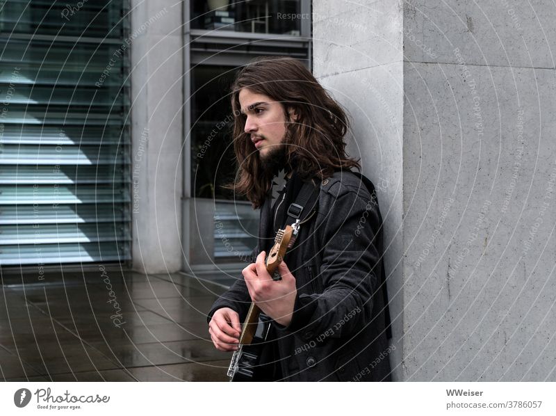 Ein junger Musiker singt im Regierungsviertel Berlin und zupft auf seiner Gitarre Bassgitarre Lied Straßenmusik nachdenklich traurig melancholisch Haare Gebäude