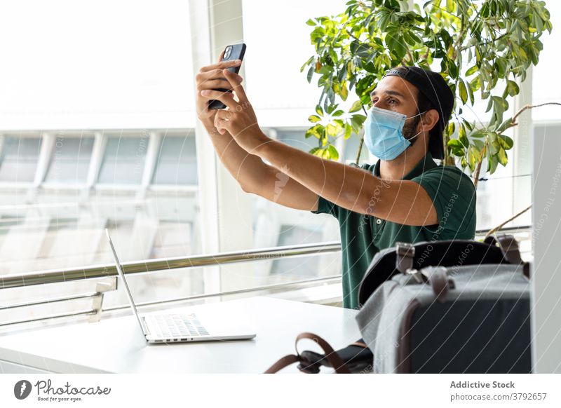 Mann mit Schutzmaske macht Selfie mit Smartphone Selbstportrait Mundschutz medizinisch Coronavirus verhindern benutzend Lächeln männlich Laptop Foto Tisch