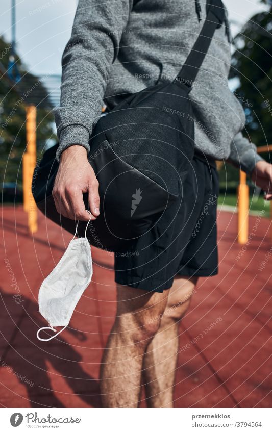 Junger Mann steht mit der Gesichtsmaske in der Hand im Calisthenics-Park auf der Strasse, um eine Virusinfektion zu vermeiden Tasche calisthenics Pflege