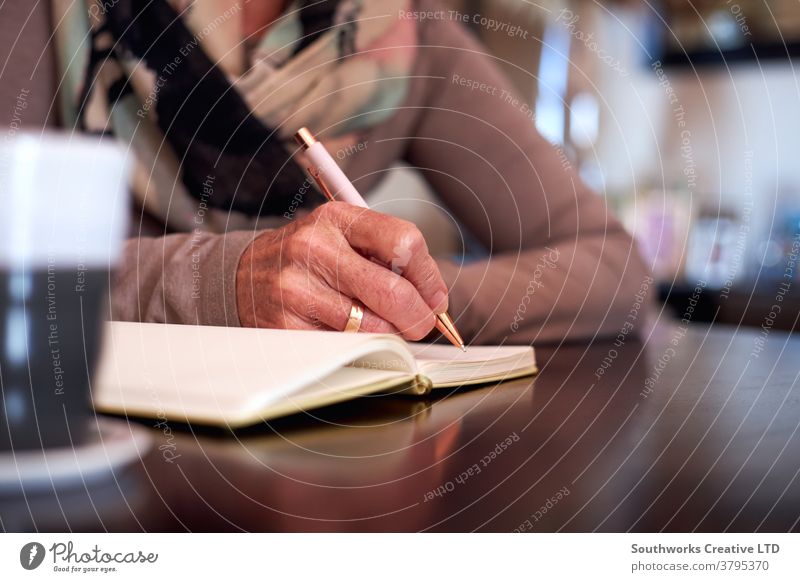 Nahaufnahme einer älteren Frau zu Hause, die am Tisch sitzt und in ein Notizbuch oder Journal schreibt Senior Senioren schreibend Tagebuch Liste erstellen