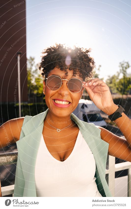 Stilvolle ethnische Frau mit Sonnenbrille in der Nähe von Zaun stehen trendy Glück Sommer Afro-Look Lächeln heiter jung Afroamerikaner schwarz positiv