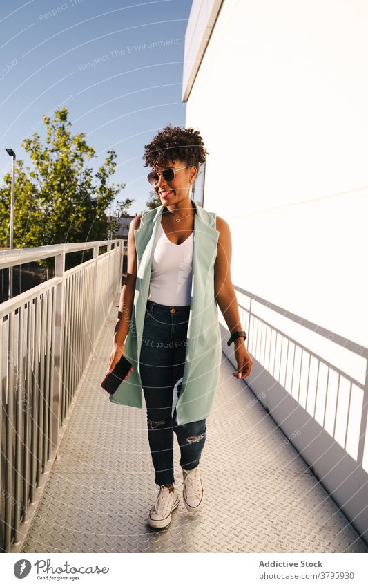 Stilvolle ethnische Frau mit Sonnenbrille in der Nähe von Zaun stehen trendy Glück Sommer Afro-Look Lächeln heiter jung Afroamerikaner schwarz positiv