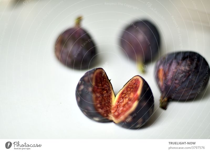 Frische, reife Feigen in dunkelblau violetter Schale, eine davon aufgeschnitten zeigt ihr rotes Fruchtfleisch, liegen auf einem weissen Tisch im hellen Tageslicht