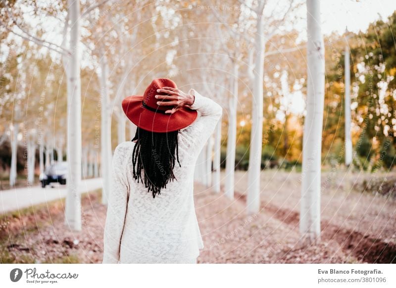 Porträt einer mittel-erwachsenen hispanischen Frau mit Hut bei Sonnenuntergang zur goldenen Stunde, Herbstzeit, schöner Baumpfad im Hintergrund. Afrofrau Latein