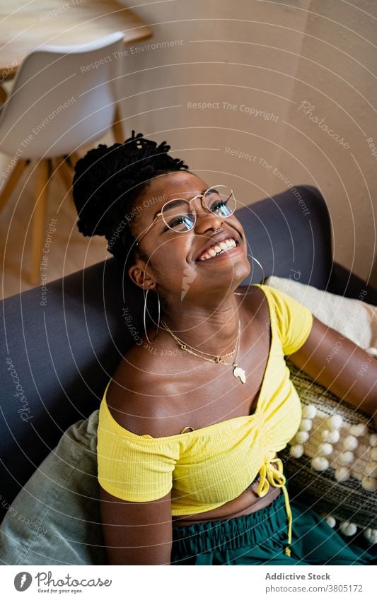 Fröhliche schwarze Frau, die sich im Wohnzimmer ausruht Zahnfarbenes Lächeln Glück zu Hause Stil Mode ruhen Wochenende Freizeit Sofa Afroamerikaner Brille