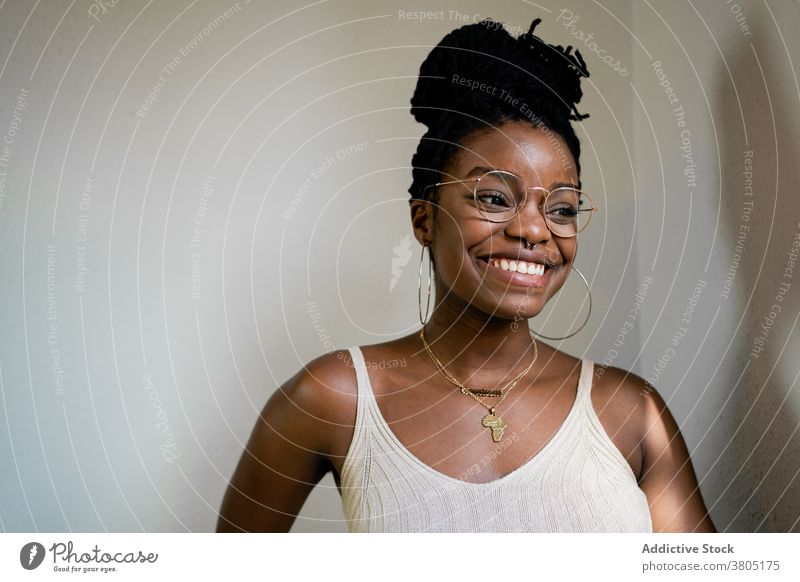 Glückliche schwarze Frau mit Brille im Zimmer Zahnfarbenes Lächeln Frisur Accessoire Mode Outfit Stil Inhalt heiter Raum Afroamerikaner brünett Freude positiv