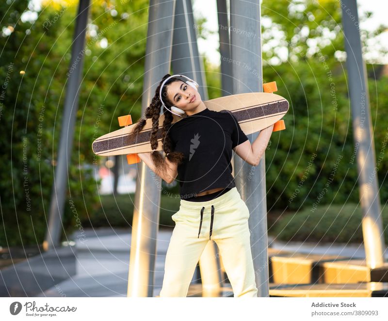 Fröhliche ethnische Dame mit Longboard genießt Musik in Kopfhörer im Park Frau zuhören Skateplatz Glück aktiv heiter trendy Baum Training modern jung