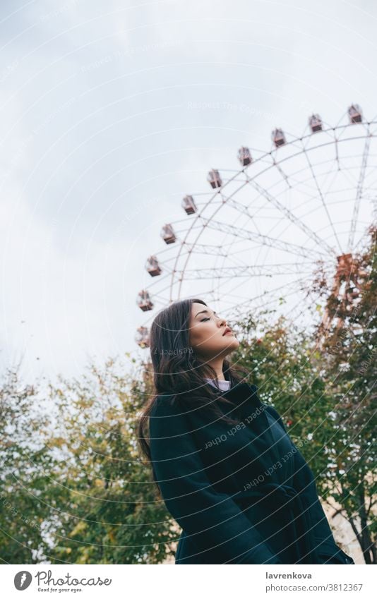 Junge erwachsene Asiatin in dunklem Mantel vor dem Riesenrad im Freien, selektiver Fokus Frau asiatisch Herbst Park lässig Freizeit saisonbedingt Wochenende