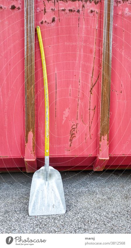 Nach getaner Arbeit lehnte die Aluschaufel mit gelbem, durchgebogenem Stil, entspannt an den roten, rostigen Stahlcontainer Schaufel Container knallig