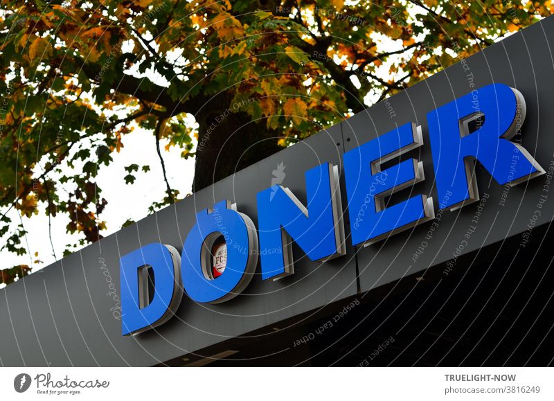 DÖNER im Herbst. Königsblauer Schriftzug DÖNER an Dach Fassade eines Schnell-Imbiss Kiosk schräg vor herbstlich gefärbtem Baum Döner Display Werbung Reklame