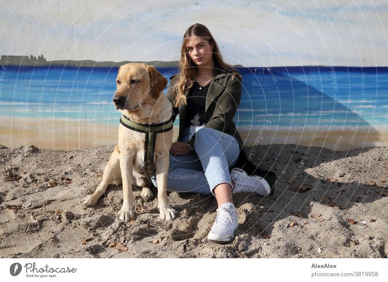 Blondes Mädchen und eine blonder Labrador sitzen an einer Wand mit Graffiti am Strand Landschaft intensiv jugendlich freundlich Natur weiblich einzigartig