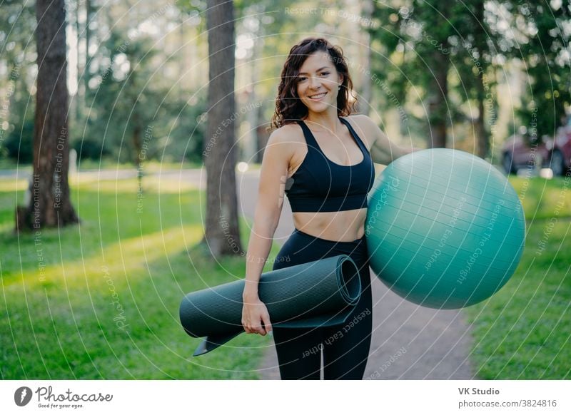 Sportliche Europäerin in kurz geschnittenem Top und Leggings, trägt zusammengerollte Karematte und Fitnessball, wird Aerobic-Übungen machen, posiert im Freien vor grünem Naturhintergrund, hat Pilattkurs