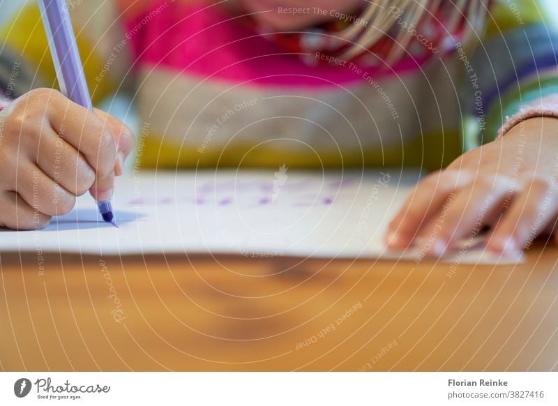 Ein 4-jähriges blondes Mädchen mit einem bunt gestreiften Pullover sitzt an einem Holztisch und zeichnet mit einem violetten Bleistift auf ein weißes Blatt Papier