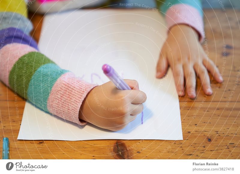 Ein 4-jähriges blondes Mädchen mit einem bunt gestreiften Pullover sitzt an einem Holztisch und zeichnet mit einem violetten Bleistift auf ein weißes Blatt Papier