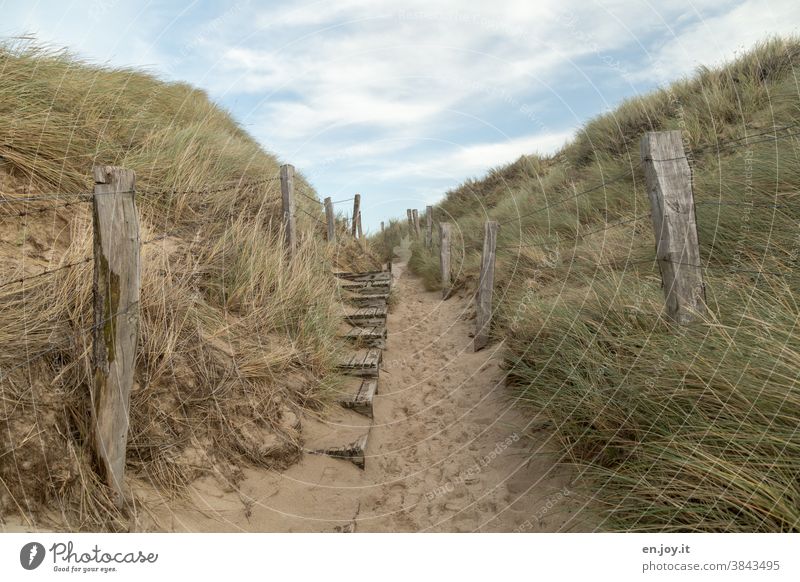 Alte und kaputte Holztreppe und Pfosten  mit Stacheldraht Zaunzwischen zwei Dünen mit Dünengras beim Übergang zum Sand Strand mit vielen Spuren unter blauem Himmel mit Wolken