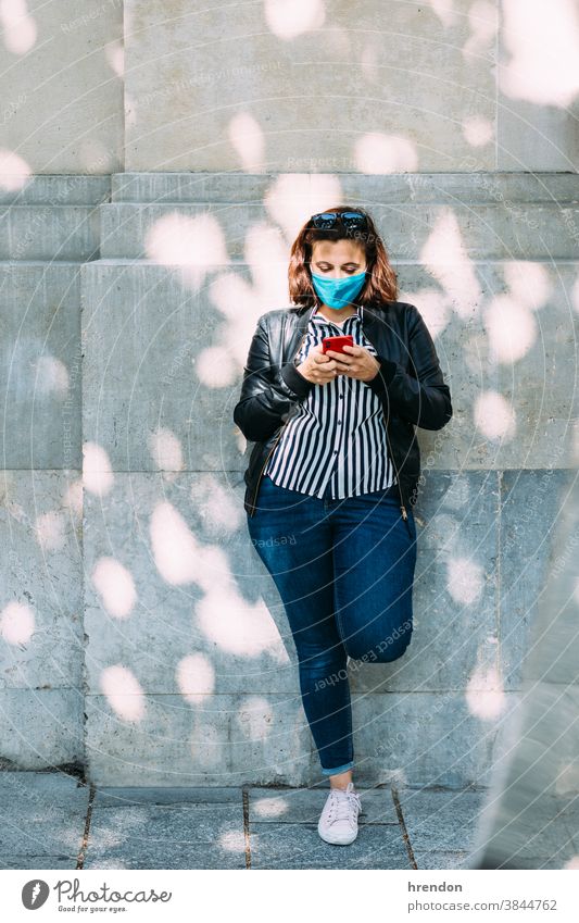 Frau mit einer Gesichtsmaske, die mit ihrem Smartphone an der Wand lehnt jung Telefon Lifestyle Lehnen Internet Mitteilung Technik & Technologie Mobile