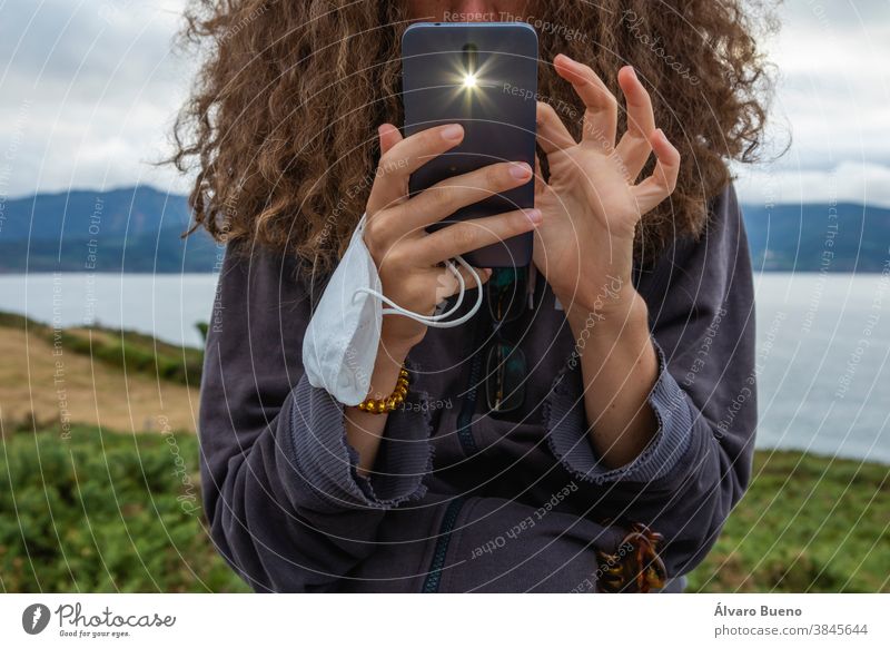 Eine junge Frau, die nach einer Gesichtsmaske und einem intelligenten Mobiltelefon greift, Spanien. Telefon Mädchen Behaarung lockig schützend Mundschutz neu