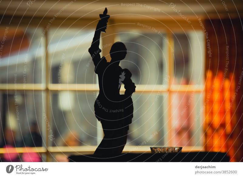 Lichtgestalt im Schatten. Figur aus Metall steht auf einem Tisch. Im Hintergrund ein Fenster und rechts eine bunte Gardine , gemustert. gestallt schwarz dunkel