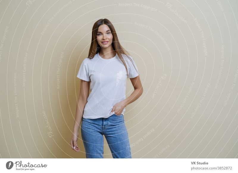 Studioaufnahme einer hübschen, schlanken Frau, die die Hand in der Tasche hält, lässiges T-Shirt und Jeans trägt, in entspannter Pose steht, selbstbewusst in die Kamera schaut, isoliert über beigen Hintergrund Gespräche mit dem Kunden führt