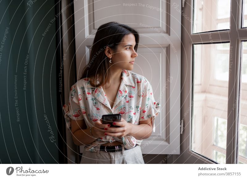 junge Frau mit Smartphone Fenster Appartement Moment Windstille Apparatur Gerät Mobile Internet benutzend Text Browsen stehen heimwärts Talkrunde brünett flach