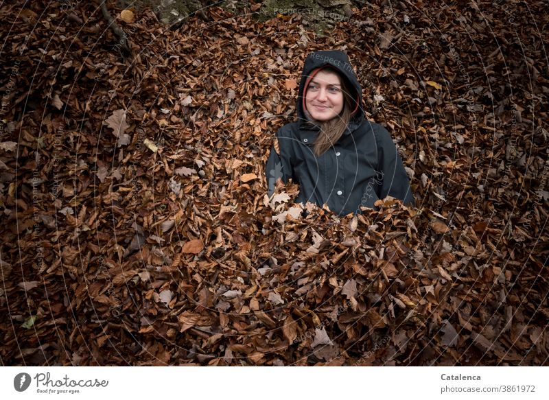 Junge, lächelnde Frau im Laub Junge Frau sitzen lange Haare brünett Blätter Pflanze Flora Herbst Anorak Schwarz Braun Blätterhaufen