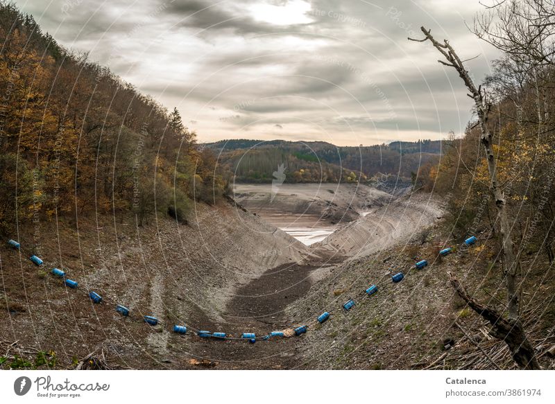 Die Begrenzung aus blauen Plastiktonnen in dem entleerten Stausee im Herbst erreicht ihren Tiefpunkt Grau Wolken Himmel herbstlich Steine Lehm Urft Fluß