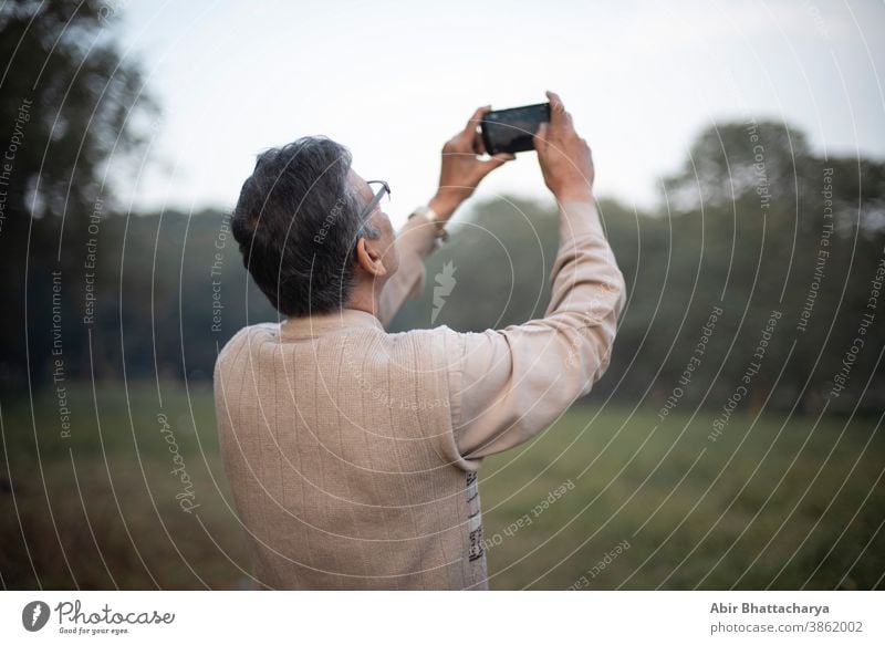 Verso Portrait eines alten indischen Bengalis im Wollpullover, der am Winternachmittag in einem Feld vor grünem Hintergrund mit seinem Mobiltelefon Himmel und Bäume fotografiert. Indischer Lebensstil und Winter.