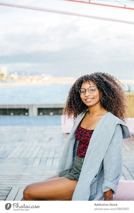 Fröhliche schwarze Frau sitzt auf Böschung Stauanlage MEER Lächeln Stil Wochenende Glück Outfit ruhen jung Kai heiter ethnisch Afroamerikaner sitzen Wasser