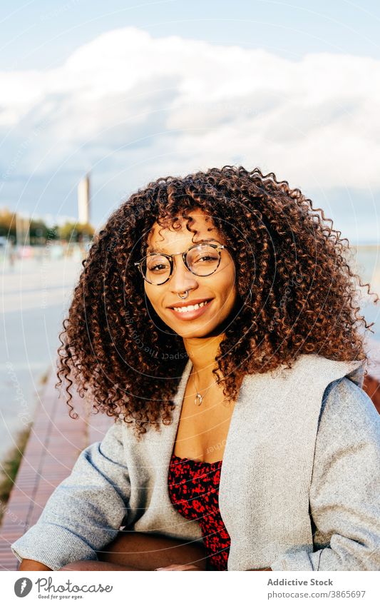 Fröhliche schwarze Frau sitzt auf Böschung Stauanlage MEER Lächeln Stil Wochenende Glück Outfit ruhen jung Kai heiter ethnisch Afroamerikaner sitzen Wasser