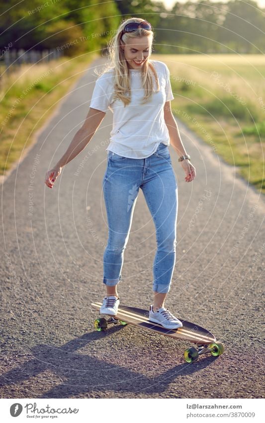 Passgenaue aktive Frau mittleren Alters spielt auf ihrem Skateboard und nähert sich der Kamera entlang einer schmalen Landstraße mit einem glücklichen Lächeln im Gegenlicht der Abendsonne