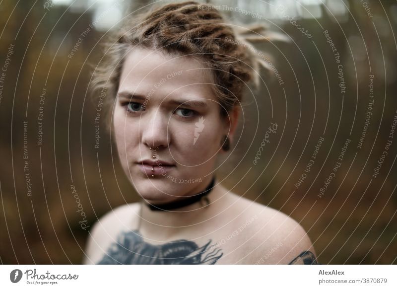 Portrait einer jungen Frau mit Dreadlocks und Tätowierungen im Wald dunkelblond Schmuck Piercing Ohrring Dekolleté Oberkörper nackt verdeckt direkt nahe Haut