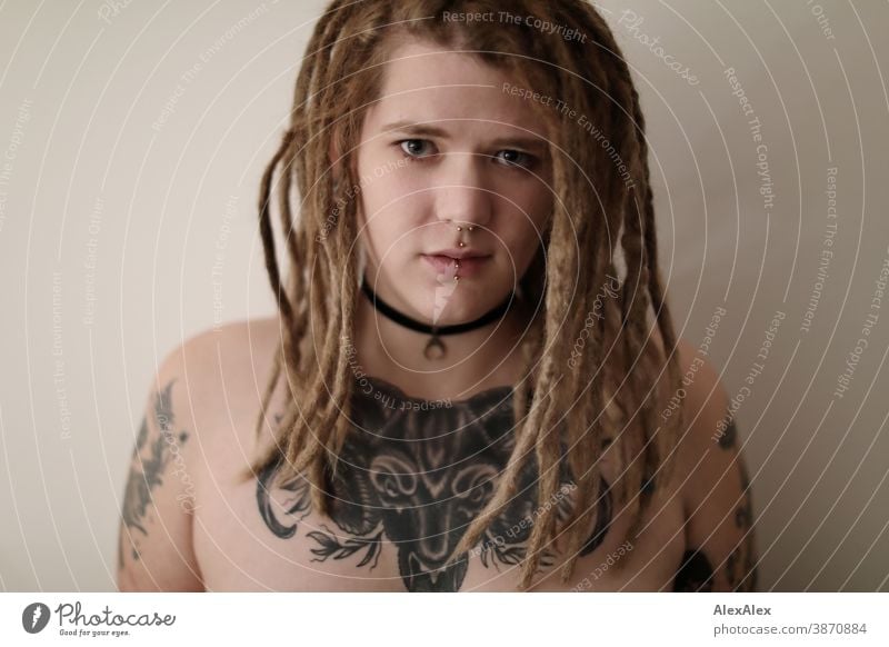 Portrait einer jungen Frau mit Dreadlocks, die eine großflächige Tätowierung eines Ochsenkopfes auf den Dekolletè hat dunkelblond Schmuck Piercing Ohrring