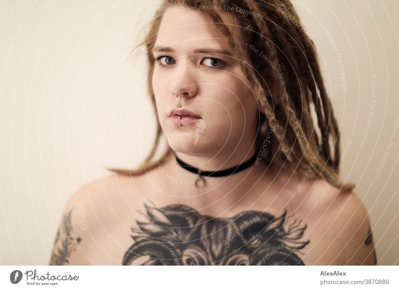 Portrait einer jungen Frau mit Dreadlocks, die eine großflächige Tätowierung eines Ochsenkopfes auf den Dekolletè hat dunkelblond Schmuck Piercing Ohrring