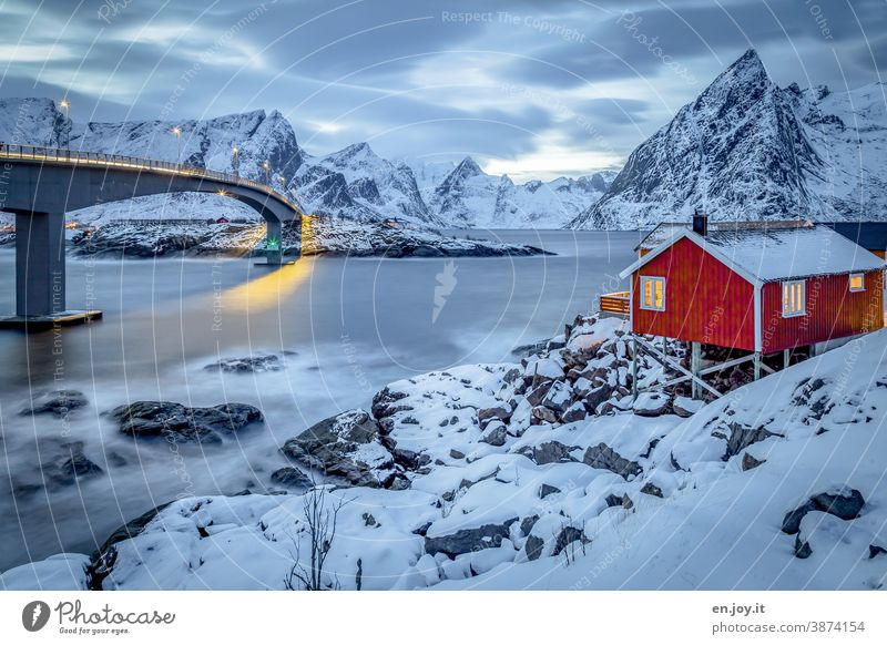 Rotes Holzhaus mit Blick auf den Reinefjord und Brücke im Winter mit viel Schnee zur blauen Stunde Lofoten Hamnøy Norwegen Skandinavien Norden Hütte Ferienhaus