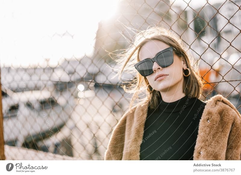 Stilvolle Frau in warmer Jacke in der Stadt Fell trendy Outfit Bestimmen Sie Sonnenbrille Oberbekleidung Netz Zaun Straße urban cool Mode ernst Windstille