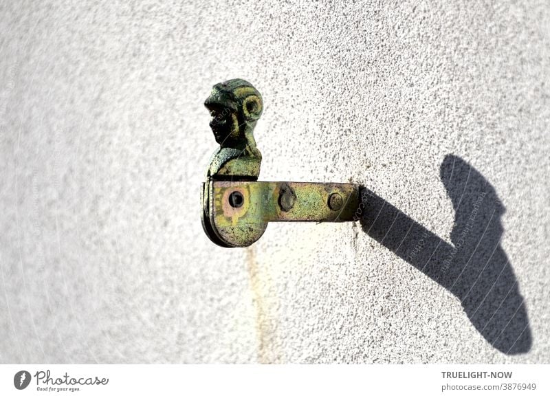 Verzinkter Fensterladenhalter  in Form eines Ritterkopfes oder Sturmhaubenträgers mit Drehgelenk und zwei Nieten im Träger ist in eine weisse Hauswand eingemauert und wirft im Sonnenlicht einen starken Schatten