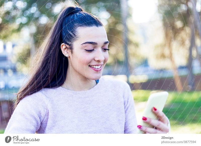 Junges, schönes, lächelndes Mädchen sitzt im Park und benutzt ihr Mobiltelefon jung Frau Telefon Sitzen Mobile Lächeln Technik & Technologie Smartphone