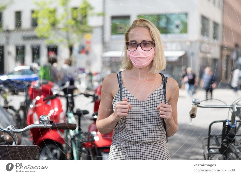 Porträt einer jungen Frau, die mit einer Schutzmaske zum Schutz vor dem Covid-19-Virus auf der Straße geht. Beiläufige Personen im Hintergrund Mundschutz