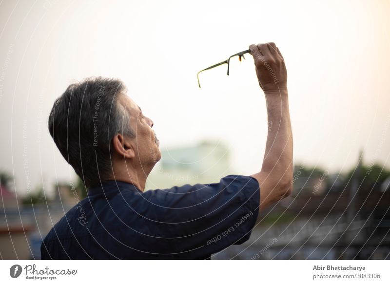 Ein alter indisch-bengalischer Mann in blauem Hemd hält seine Brille in die Luft, um sie aufmerksam zu beobachten, während er auf einem Dach unter freiem Himmel steht. Indischer Lebensstil und Senioren