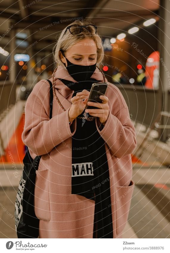 Junge Frau steht am Bahnhof mit einer Koronamaske Coronavirus blond lange Haare Reise Unternehmen Winter Mantel Brille Mode Stil Blick hübsch Porträt Farbfoto