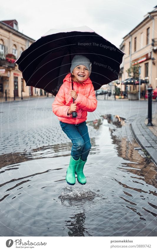 Kleines Mädchen mit großem Regenschirm in der Hand springt an einem verregneten, düsteren Herbsttag in die Pfütze im Freien wenig saisonbedingt fallen Kindheit