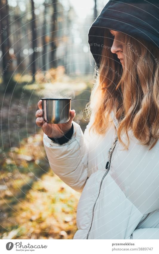 Frau mit Kapuze, die während einer Herbstreise Pause macht und am kalten Herbsttag eine Tasse mit heißem Getränk aus der Thermoskanne hält aktiv Aktivität