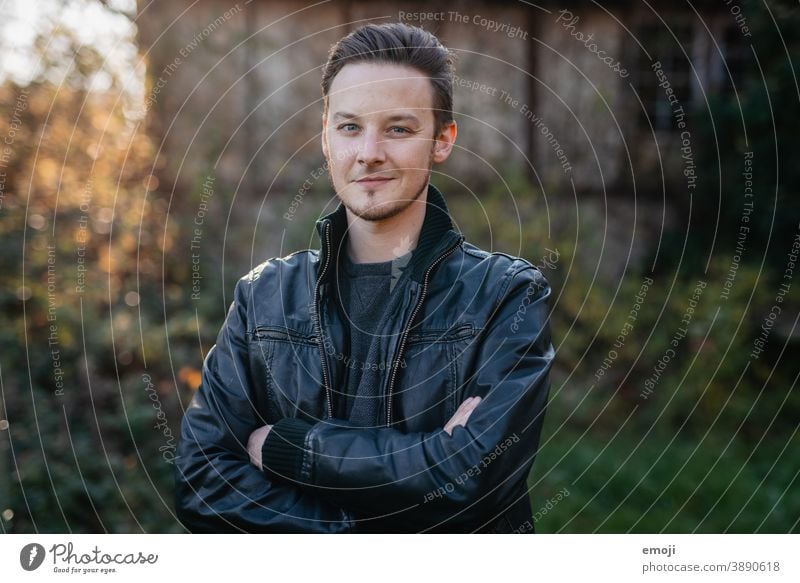 junger Mann in Lederjacke draussen im Freien grün männlich Erwachsener Coolness Oberkörper die Arme verschränkt Junger Mann Freundlichkeit Lächeln positiv