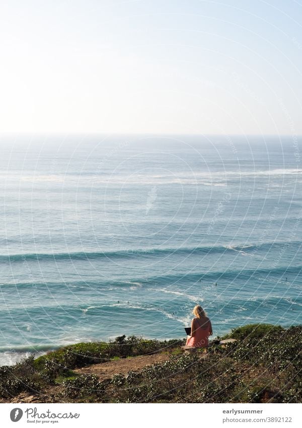 Frau sitzt auf einer Klippe und arbeitet an ihrem Laptop mit Blick auf den Ozean und die Wellen in Portugal Remote-Job digitaler Nomade Selbstständigkeit