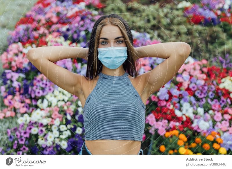 Junge Frau in medizinischen Maske stehen in der Nähe von blühenden Blumen Mundschutz behüten Park Coronavirus Bund 19 Pandemie neue Normale jung lässig