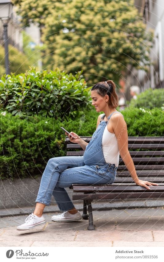 Lächelnde schwangere Frau mit Smartphone in der Stadt Großstadt Browsen Straße Bank hölzern Apparatur Windstille Surfen Anschluss online Kommunizieren Unterleib