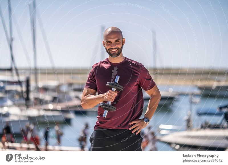 Ein Mann steht mit Gewichten im Freien am Meer Porträt Übung Sport Seeküste Park urban Tag sonnig jung stehen Textfreiraum Jachthafen Boot ernst Trainerin