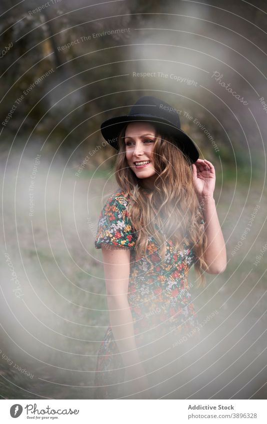 Fröhliche Frau mit Hut im Wald stehend Accessoire charmant Kleid Lächeln Outfit Natur Bekleidung Wälder Vorschein lange Haare gewelltes Haar Optimist Stil