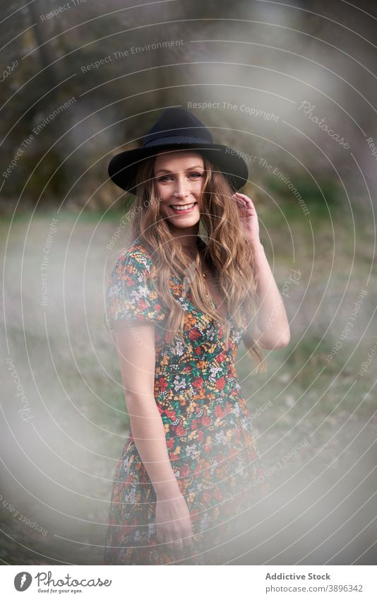 Fröhliche Frau mit Hut im Wald stehend Accessoire charmant Kleid Lächeln Outfit Natur Bekleidung Wälder Vorschein lange Haare gewelltes Haar Optimist Stil