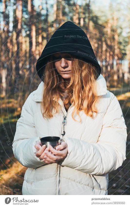 Frau mit Kapuze, die während einer Herbstreise Pause macht und am kalten Herbsttag eine Tasse mit heißem Getränk aus der Thermoskanne hält aktiv Aktivität